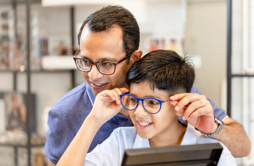 A child trying on blue glasses with guidance from a smiling adult wearing glasses, inside a brightly lit optometry clinic.